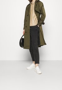 Manteau trench olive, chemise beige à boutons, pantalon noir ajusté, baskets blanches, sac noir à la main, textures lisses, design décontracté.