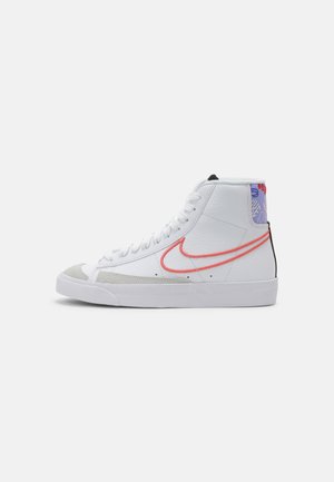 Białe wysokie trampki z czerwonym logo Nike, szarą zamszową osłoną na palce i fioletowym wzorzystym kołnierzem na pięcie na białym tle.
