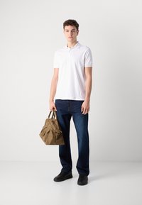 Hvitt polo skjorte, mørkeblå jeans, svarte sko, holder en brun skulderveske med flere lommer og håndtak. Enkel bakgrunn.