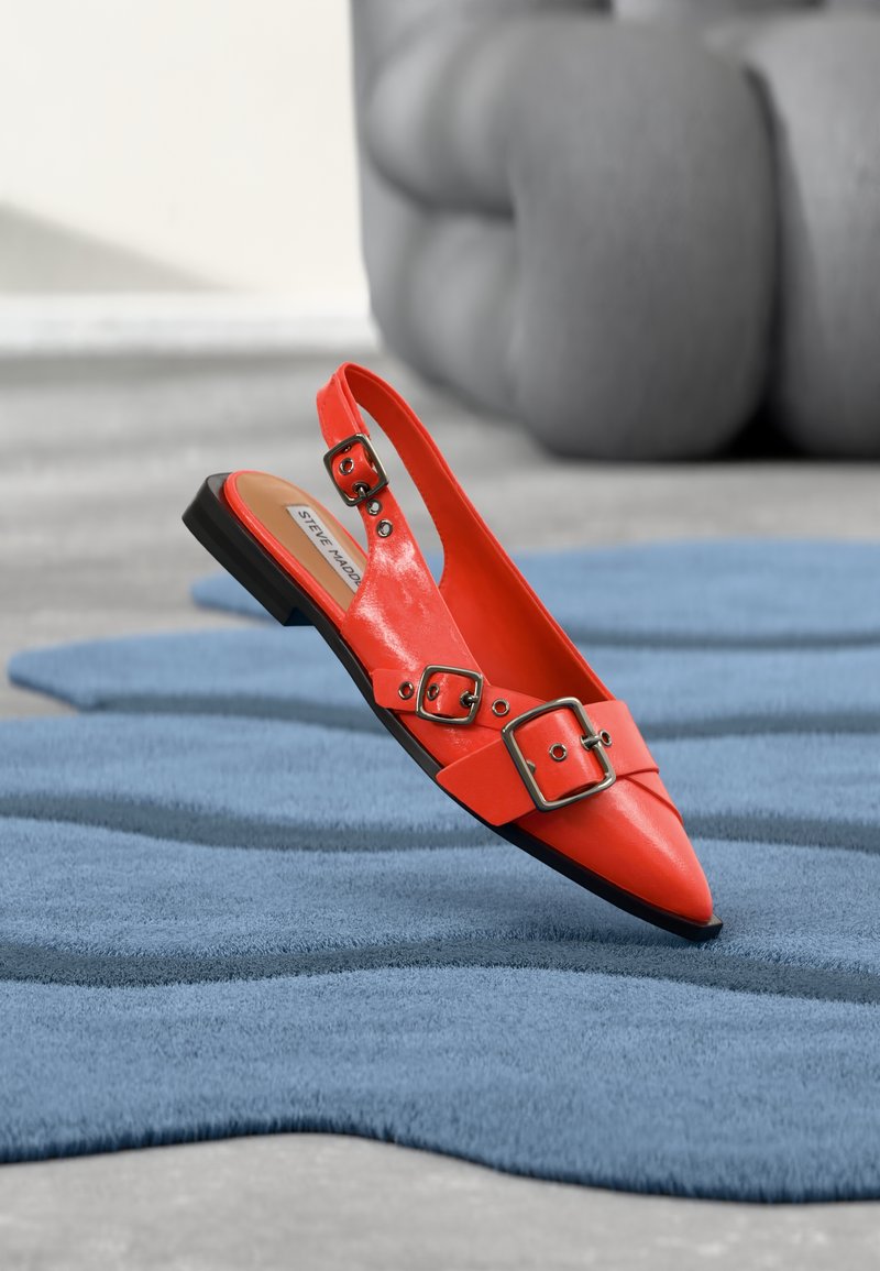 Žiarivo oranžová plochá slingback obuv so špicatou špičkou a tromi prackami, položená na textúrovanom modrom koberci v modernom priestore.