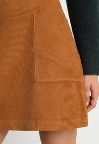 Hnědá kordurová sukně s drsnou texturou, boční kapsou a rovným střihem. Materiál se zdá být odolný a teplý.