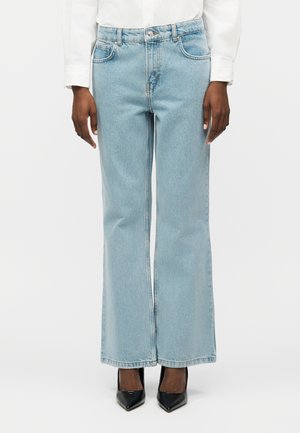 Osoba oblečená v svetlomodrých džínsoch s vysokým pásom, širokými nohavicami a prednými vreckami, a čiernych topánkach s ostrou špičkou, v kombinácii s bielou košeľou s dlhým rukávom.