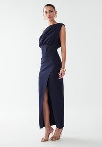 Vestido azul marino de un solo hombro con un corpiño ajustado, detalles fruncidos, una abertura lateral y tela suave. Combinado con tacones de tiras de color claro.