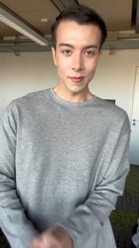 Persoană tânără cu păr scurt și închis la culoare, purtând un tricou gri cu mâneci lungi, stând într-un spațiu modern de birou interior.