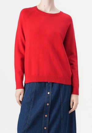 Kvinde iført en knald rød langærmet striktrøje kombineret med et mørkeblåt denimskørt med knapper foran.