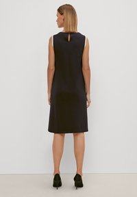 Ärmelloses schwarzes Kleid mit glatter Textur, knielangem Design und einem Schlüssellochdetail am Rücken, kombiniert mit schwarzen High Heels.