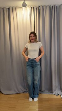 Femme debout à l’intérieur les mains dans les poches, portant un t-shirt blanc, un jean bleu et des chaussettes blanches, avec des rideaux gris fermés et un sol en bois en arrière-plan.