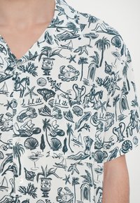 Κοντό μανίκι μπλούζα από λευκό ύφασμα με μαύρη τροπική εκτύπωση που περιλαμβάνει διάφορες θαλάσσιες ζωές, φυτά και αντικείμενα. Ορατή λεπτομέρεια με κουμπί.