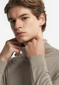 Beige polotröja med ribbade manschetter och krage. Modellen visar händer som justerar nackområdet, vilket framhäver den mjuka, släta tygtexturen.