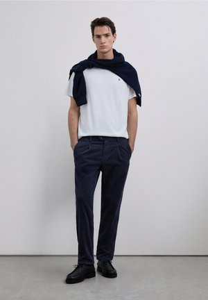 T-shirt bianca di cotone, maglione lavorato a maglia blu scuro appoggiato sulle spalle, pantaloni in velluto a coste blu scuro, scarpe nere con stringhe, in piedi contro uno sfondo neutro.