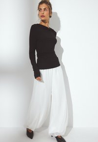 Juodas ilgomis rankovėmis viršus su susiraukšlėjimu, derinamas su plačiomis baltomis kelnėmis. Modelis vilki smailiakūnius juodus batelius, poza fone – balta.