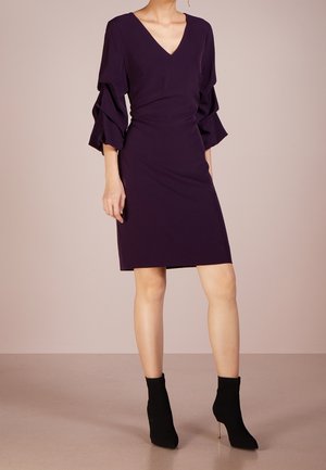 Kvinde iført en lilla knælængde kjole med rynkede trekvartærmer og sorte ankelstøvler med høje hæle, stående.