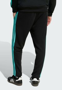 Pantalon de sport noir avec des rayures bleu sarcelle sur les côtés, coupe ajustée, taille élastique, porté avec des baskets noires et blanches.