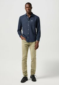 Camisa azul marino de botones con un bolsillo en el pecho, combinada con chinos ajustados de color verde claro y zapatos negros con cordones. Tejido suave y diseño estructurado.