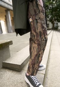 Spodnie bojówki w kamuflażu w odcieniach brązu, zieleni i beżu; posiadają kieszenie oraz naszywkę z logo; zestawione z szarymi sneakersami na platformie.