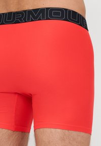 Boxer-briefs rouge en matière extensible avec une ceinture logo noire, présentant une texture lisse et un design ajusté qui s'étend jusqu'à mi-cuisse.