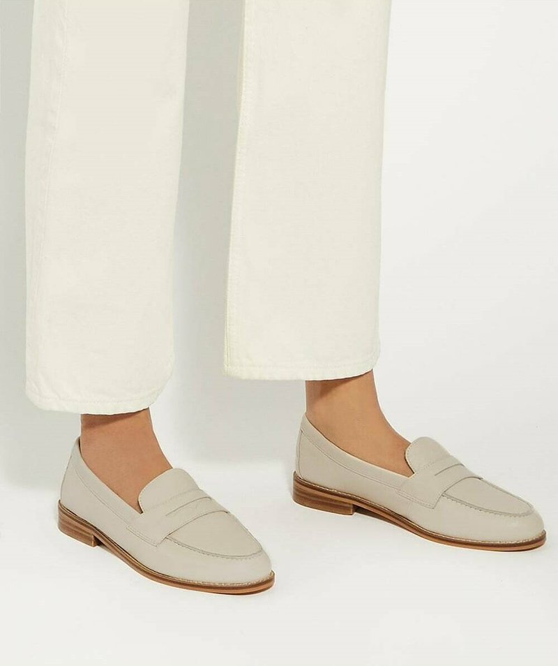 Personne portant des mocassins en cuir beige avec des semelles en bois et un pantalon blanc court sur un fond uni.