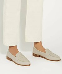 Beige leren instappers met een gladde textuur, voorzien van een gestikt ontwerp en een lage houten hak, gedragen met witte wide-leg broeken.