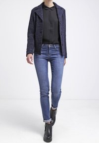 Giacca blu navy con bottoni e colletto, blusa nera sotto, abbinata a jeans skinny blu e stivaletti neri. Tessuto con texture.