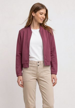 Femme portant une veste violette zippée, une chemise blanche et un pantalon beige, debout et regardant vers sa gauche sur un fond uni.
