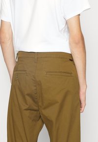 Personne portant un pantalon marron avec des poches passepoilées à l'arrière et une chemise blanche à manches courtes, vue de la taille jusqu'au milieu des cuisses, de dos.
