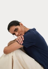 T-shirt blu navy a maniche corte realizzato in tessuto morbido, con collo rotondo e vestibilità rilassata, indossato con un tessuto chiaro e strutturato sotto.