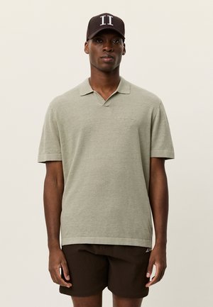 Lys grønn kortermet polo med krage, med en subtil logo. Brukt sammen med brune teksturerte shorts og en mørk caps med hvit skrift.