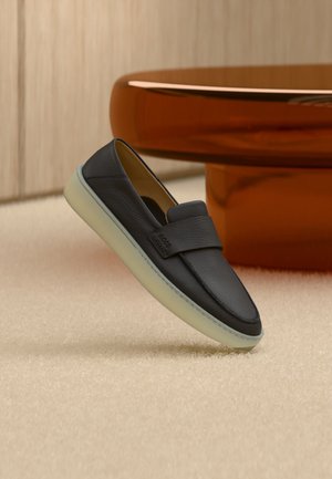 Mocassino in pelle nera con suola bianca, posato su un tappeto beige vicino alla gamba rotonda di un tavolo di legno marrone.