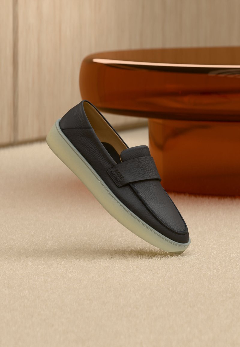 Μαύρο δερμάτινο μοκασίνι με λευκή σόλα, τοποθετημένο σε μπεζ χαλί δίπλα σε στρογγυλό καφέ ξύλινο πόδι τραπεζιού.