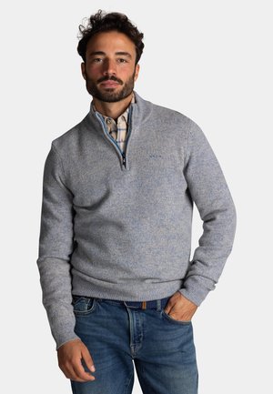 Lichtblauwe ritstrui gemaakt van een gestructureerd breimateriaal. Kenmerkt zich door ribgebreide boorden en onderkant, gedragen over een geruite geklede shirt.
