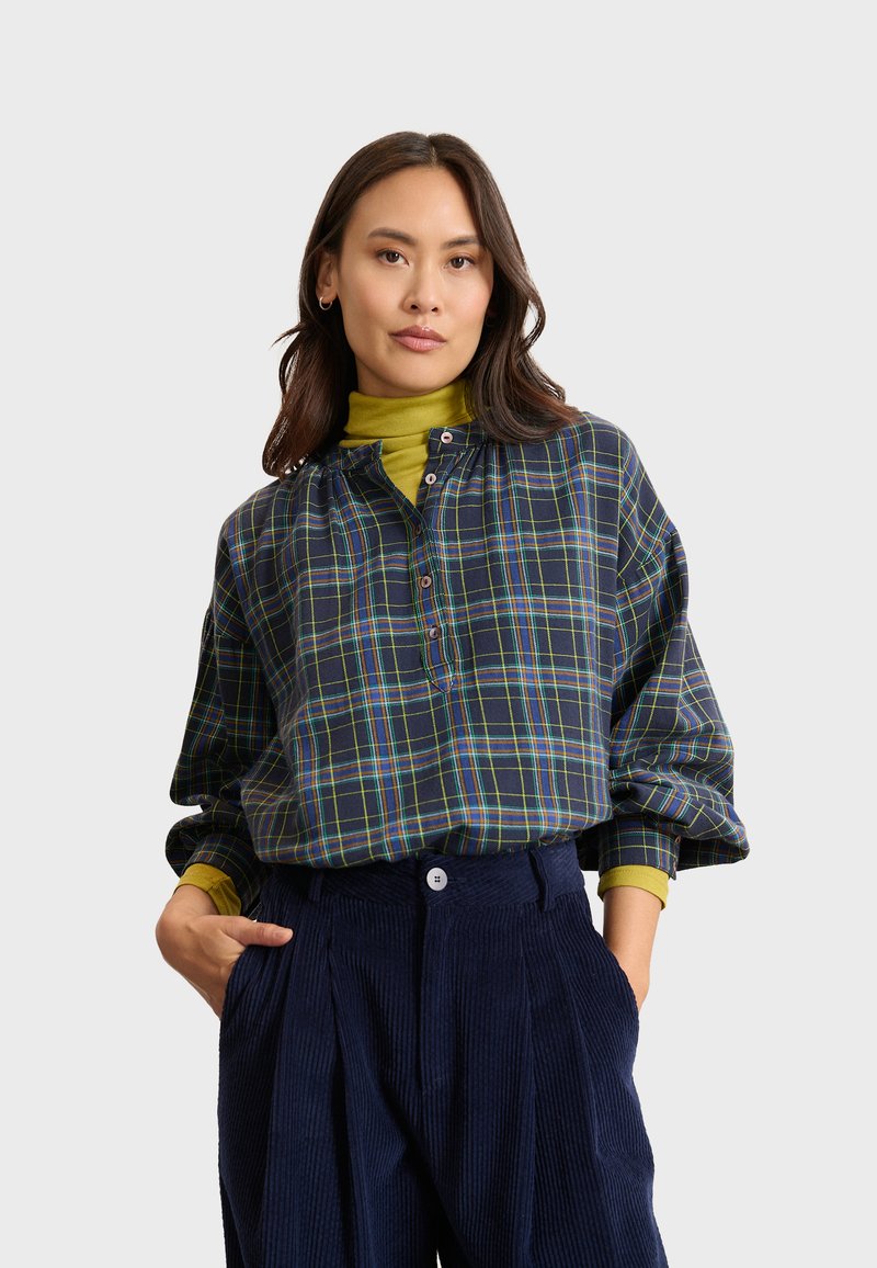 Camicia a scacchi blu navy con linee verdi, blu e gialle, dotata di colletto a bottoni e maniche lunghe, abbinata a un dolcevita verde.