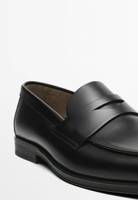 Schwarze Lederloafers mit glatter Textur, ausgestattet mit einem breiten Riemen über dem Obermaterial und einem niedrigen, gestuften Absatz. Das Innenfutter zeigt ein weiches Material.