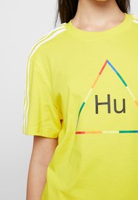 Gul bomull t-shirt med korta ärmar, med en färgglad triangeldesign och texten "Hu" i fet svart. Tre vita ränder på axlarna.