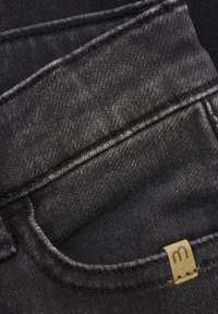 Tissu en denim noir avec une texture lisse, présentant des poches cousues et une petite étiquette avec la lettre "m" en brun.