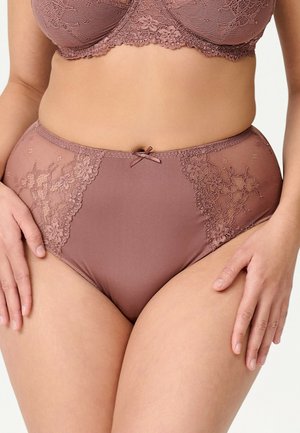 Frau trägt hoch taillierte Spitzen-Panties in Mauve und passenden BH, die Hände ruhen auf den Oberschenkeln, vor einem schlichten hellen Hintergrund.