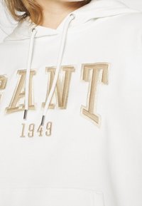Sudadera blanca con bordado en beige de la palabra "GANT" y detalle "1949". Cuenta con cordones y acentos metálicos en la capucha.