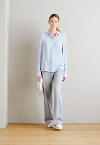 Camicia azzurro chiaro, a maniche lunghe, con bottoni e una vestibilità rilassata; abbinata a jeans grigi chiari a trombetta. La modella tiene una borsa rotonda bianca, in piedi su un pavimento di legno.
