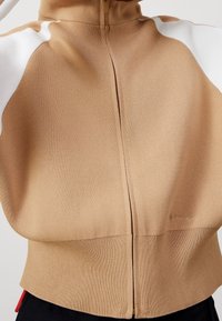 Brązowy sweter zapinany na zamek z białymi akcentami, o prążkowanej fakturze, wysokim kołnierzu i dopasowanym pasie. Dyskretna detal z logo na dolnej krawędzi.