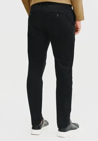 Czarne spodnie wykonane z fakturowanej tkaniny, w slim fit, z tylnymi kieszeniami zapinanymi na guziki, noszone z czarnymi trampkami i brązową koszulą.