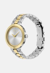 Sidabrinė ir auksinė nerūdijančio plieno laikrodžio suapvalinta korpuso forma, balta tekstūruota ciferblatas, auksiniai akcentai ir grandinėlė su pakaitiniais spalvų elementais.