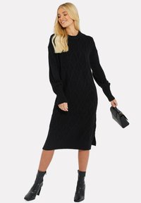 Schwarzes Strickkleid mit einem Diamantmuster, langen Ärmeln, gerippten Bündchen und Seitenschlitzen; kombiniert mit knöchelhohen Stiefeln und einer strukturierten Handtasche.