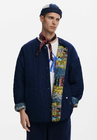 Giacca trapuntata blu navy con una fodera dal motivo colorato. Chiusure a scatto, colletto rotondo e un tessuto texturizzato. Indossata con una maglietta bianca.