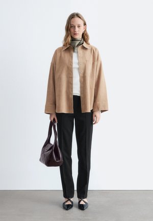 Nainen, joka käyttää beigeä ylipitkää paitaa, valkoista toppia, mustia housuja, mustia piikkikärkisiä kenkiä, vihreää pallokuvioista huivia ja pitää kädessään tummanruskeaa nahkalaukkua.