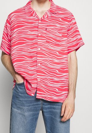 Personne portant une chemise rouge à manches courtes avec des rayures blanches ondulées et un jean bleu, main dans la poche, sur un fond uni.