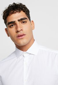 Camisa blanca de botones con cuello puntiagudo, que presenta una textura suave y un diseño clásico. El modelo exhibe una expresión neutral y tiene el cabello corto y rizado.