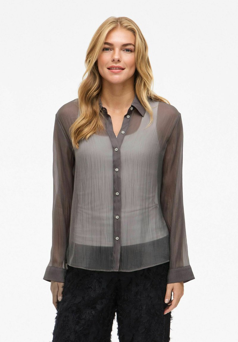 Transparante grijze blouse met een verticaal geplooid patroon, knoopsluiting aan de voorkant en lange mouwen; heeft een subtiele glans en een zachte textuur.