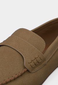 Καφέ μοκασίνι από σουέτ με ραφές μοκασίνου και διακοσμητικό λουράκι πάνω από το επάνω μέρος, σε κοντινή προβολή.