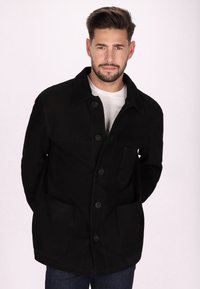 Veste noire à boutons avec un col, deux poches avant et une texture douce. Portée par-dessus une chemise blanche, présentant une coupe droite.