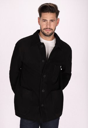 Veste noire à boutons avec un col, deux poches avant et une texture douce. Portée par-dessus une chemise blanche, présentant une coupe droite.