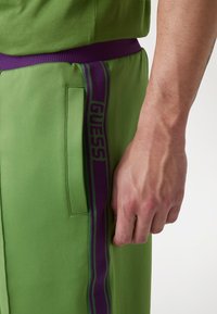 Zeleni športni hlače z vijoličnim elastičnim pasom, ki ima stransko žep in logo "GUESS" na stranskem delu, gladka tekstura.